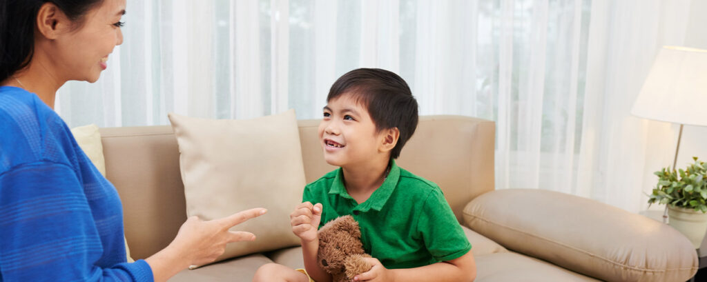 Boy sits on couch, hugs teddy bear, and talks with woman