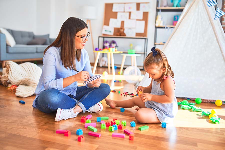 Woman sits on floor and watches child play with blocks