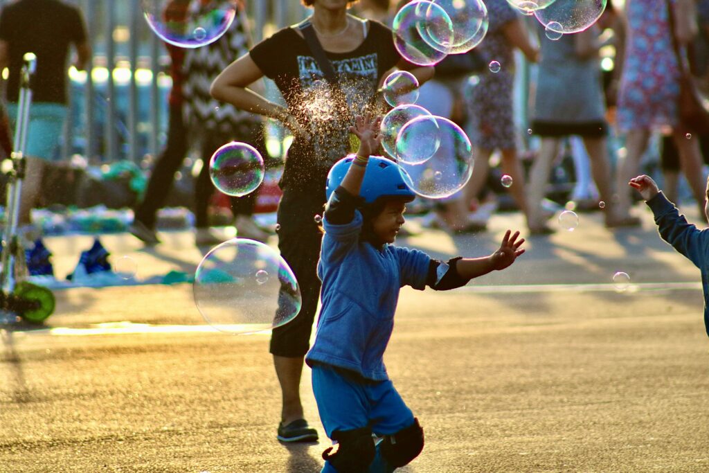 A young child wearing a blue helmet and protective pads reaches up playfully toward floating soap bubbles at an outdoor family outings community street festival on a sunny day.