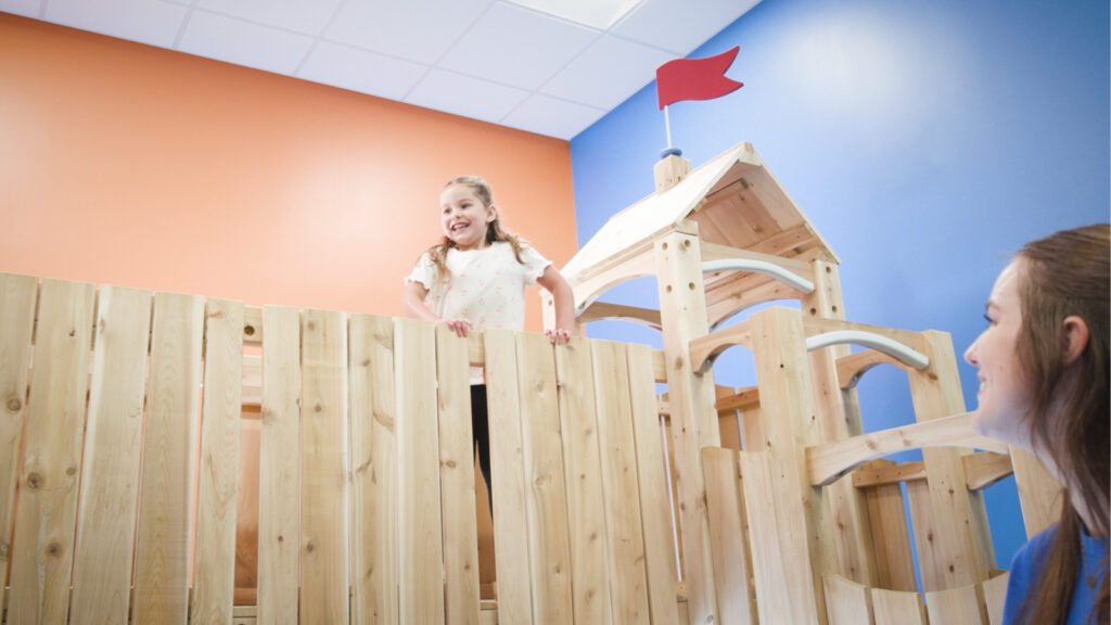 Child grinning from the top of a wooden indoor playground.