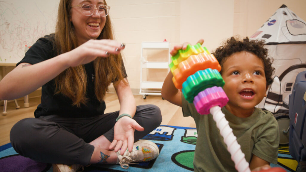 Child showing toy to someone offscreen while playing with another adult sitting on the floor.