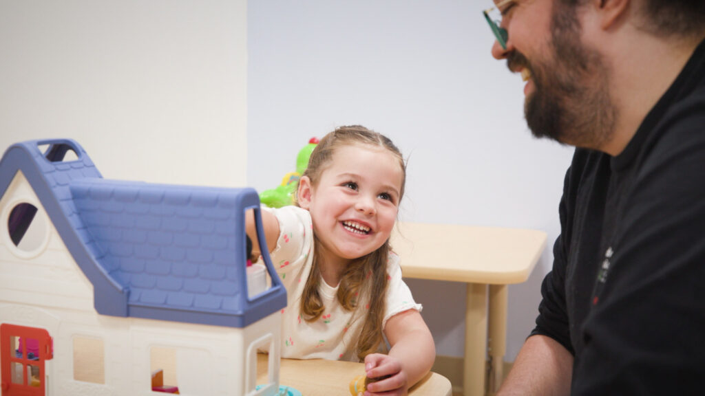 Child grinning while playing with a toy house with an adult.