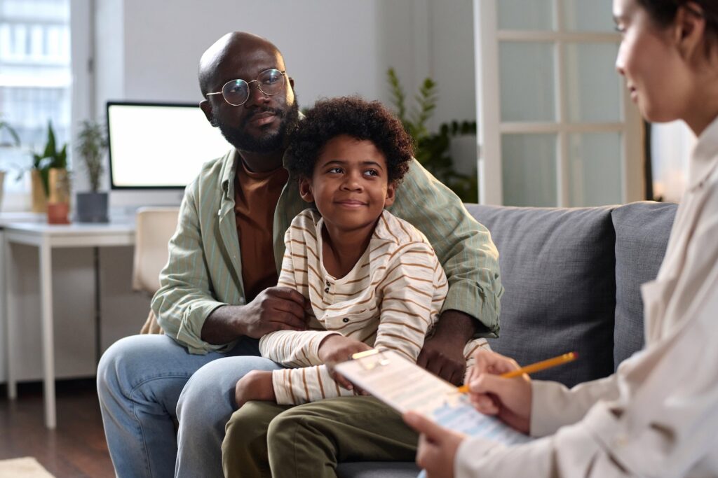 A father and young son sit together on a couch, attentively listening to a therapist who is taking notes during a counseling session.