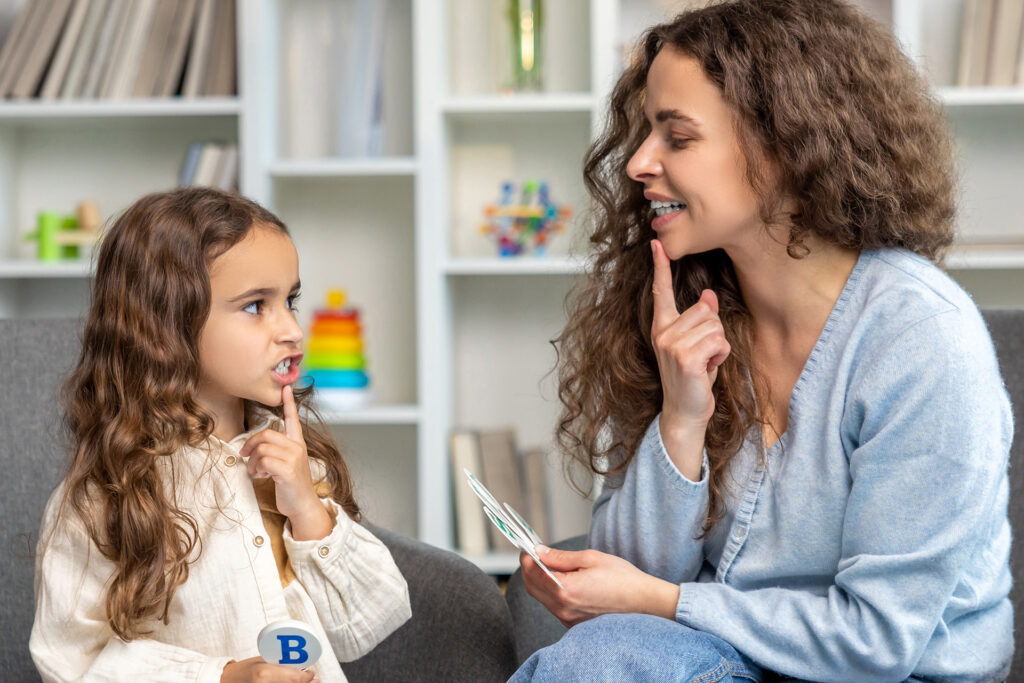 A female speech-language pathologist and a young girl sit together during a therapy session. The therapist points to her chin to demonstrate a speech sound, and the girl mirrors the gesture while holding a letter "B" block.