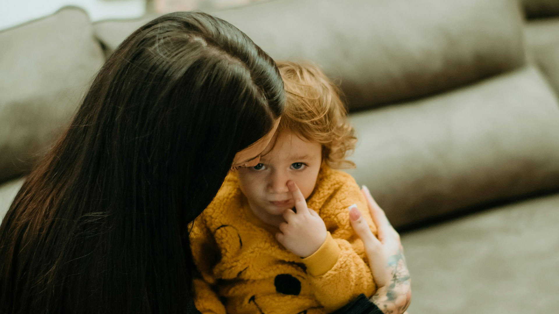 An overhead, medium shot shows a dark-haired woman holding a small child with light-colored curly hair. The woman’s back is to the camera as she leans her head toward the child. The child wears a yellow, fuzzy sweater and looks toward the camera while holding a finger to their nose. The background is a blurry grey sofa.