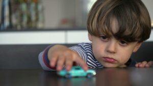 Alt Text: A young child sitting at a table with a calm expression, resting his head near his hands as he gently pushes a small toy car away.