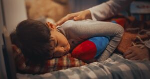 A young child asleep in a bed with plaid and striped bedding, hugging a blue and red plush toy. An adult's hand rests gently on the child's shoulder.
