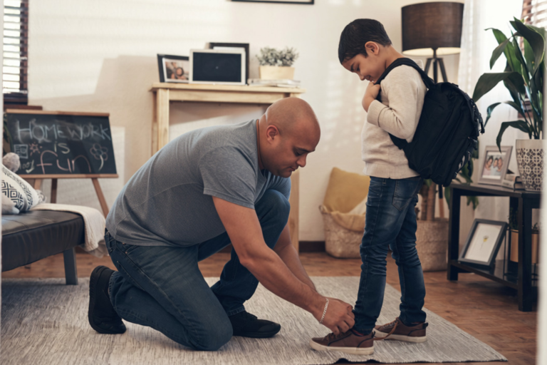 A father kneeling down on a living room rug to help his young son tie his shoes before school.