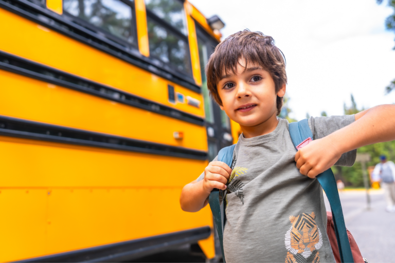 A young boy holding his backpack straps while standing in front of a bright yellow school bus.