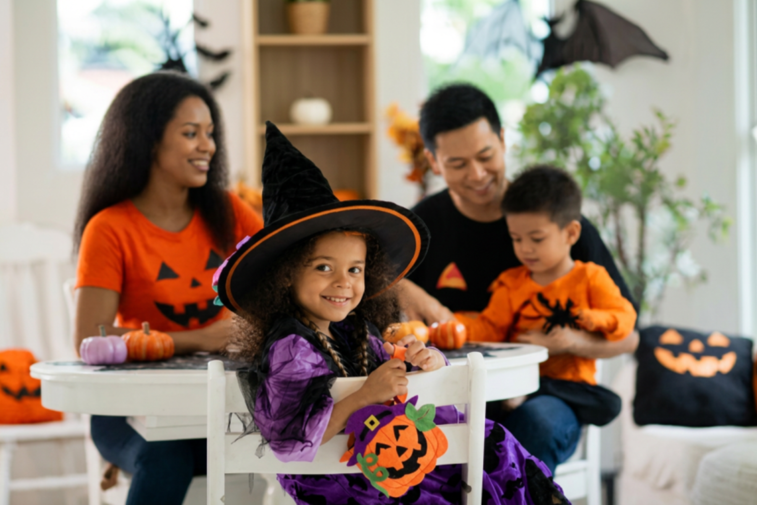 A young girl dressed in a purple witch costume and hat sits in the foreground at a table, with her smiling family in Halloween-themed clothing in the background.
