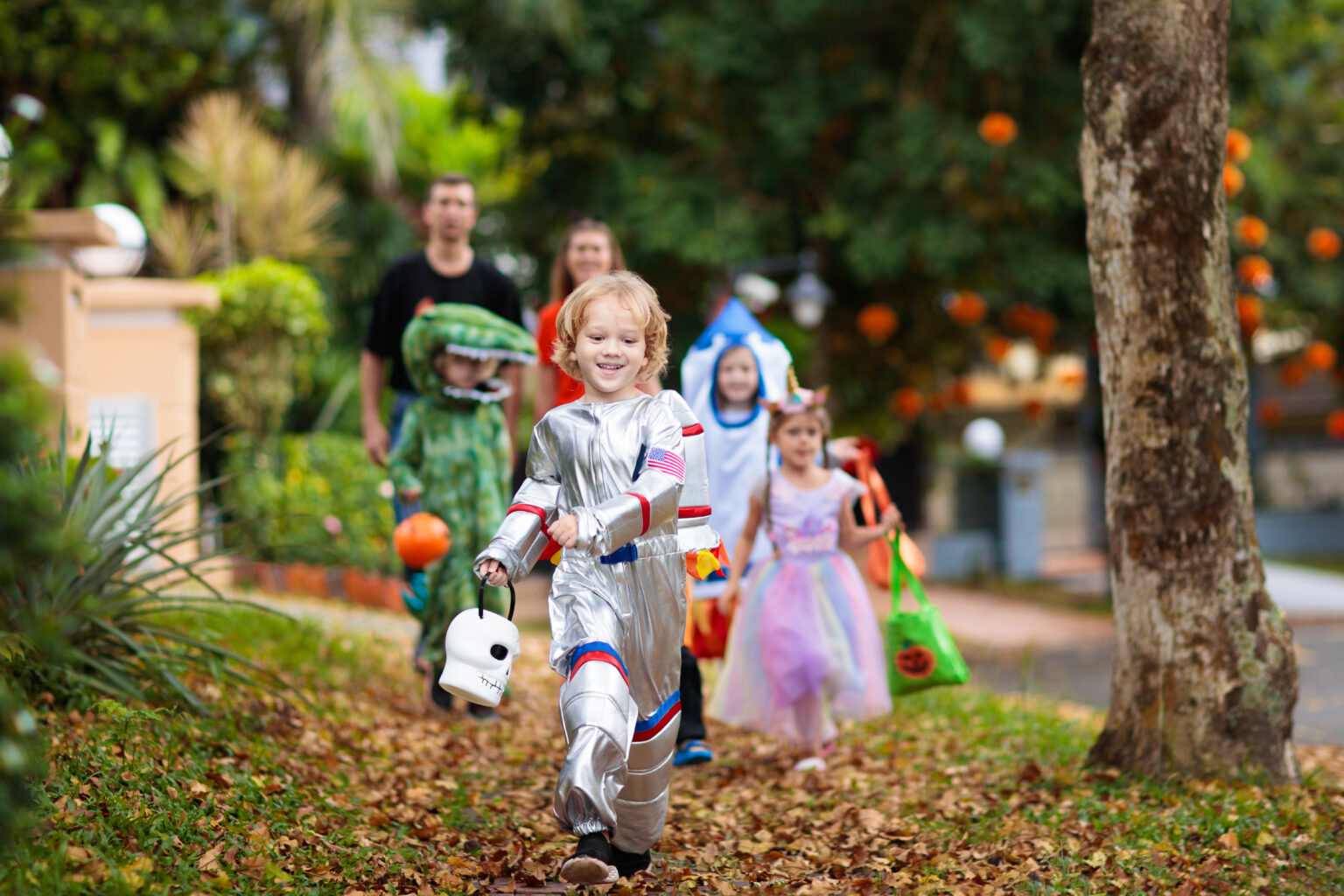 Children in costumes, including an astronaut and a dinosaur, walking along a neighborhood path while trick-or-treating for Halloween safety tips illustration.