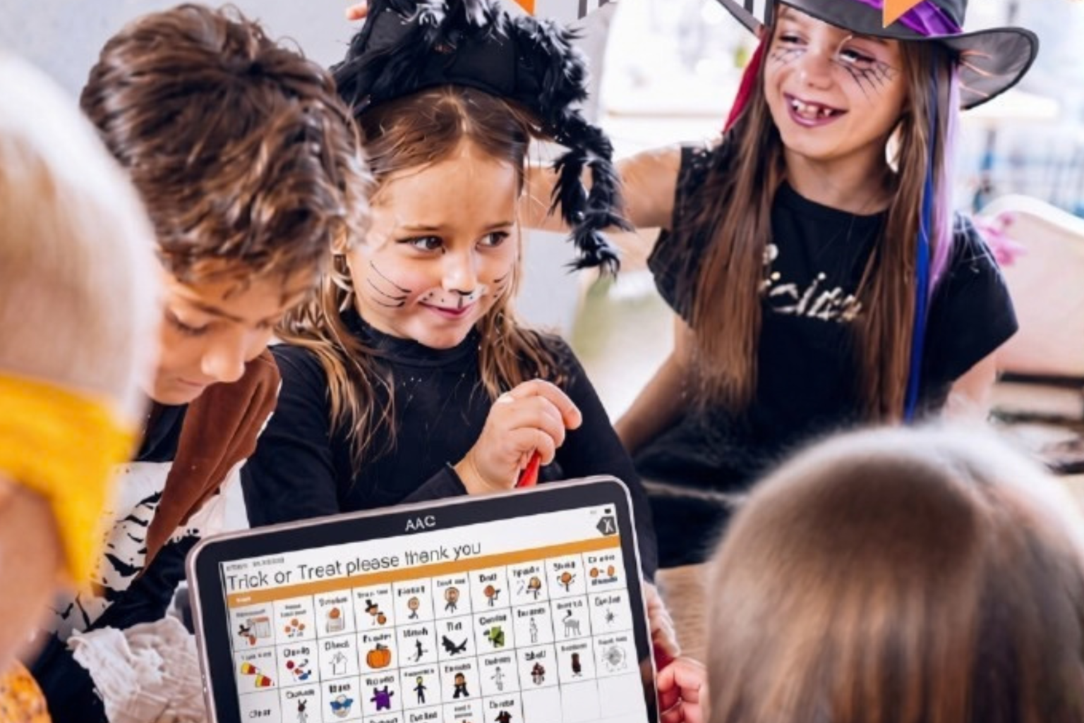 A group of young children in Halloween costumes gathered around a tablet displaying a custom AAC speech screen with trick-or-treat picture symbols.