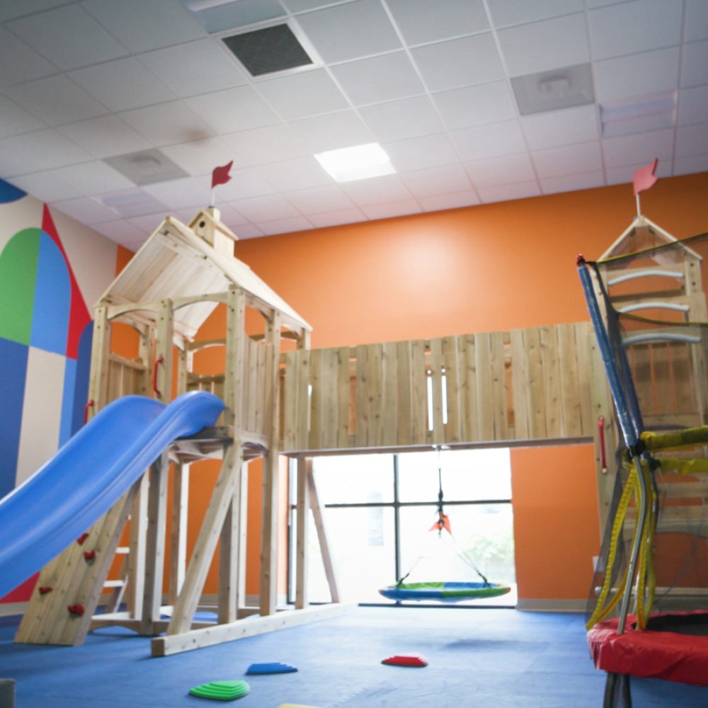 Indoor playground including a wooden jungle gym with a slide and bridge, and a trampoline.